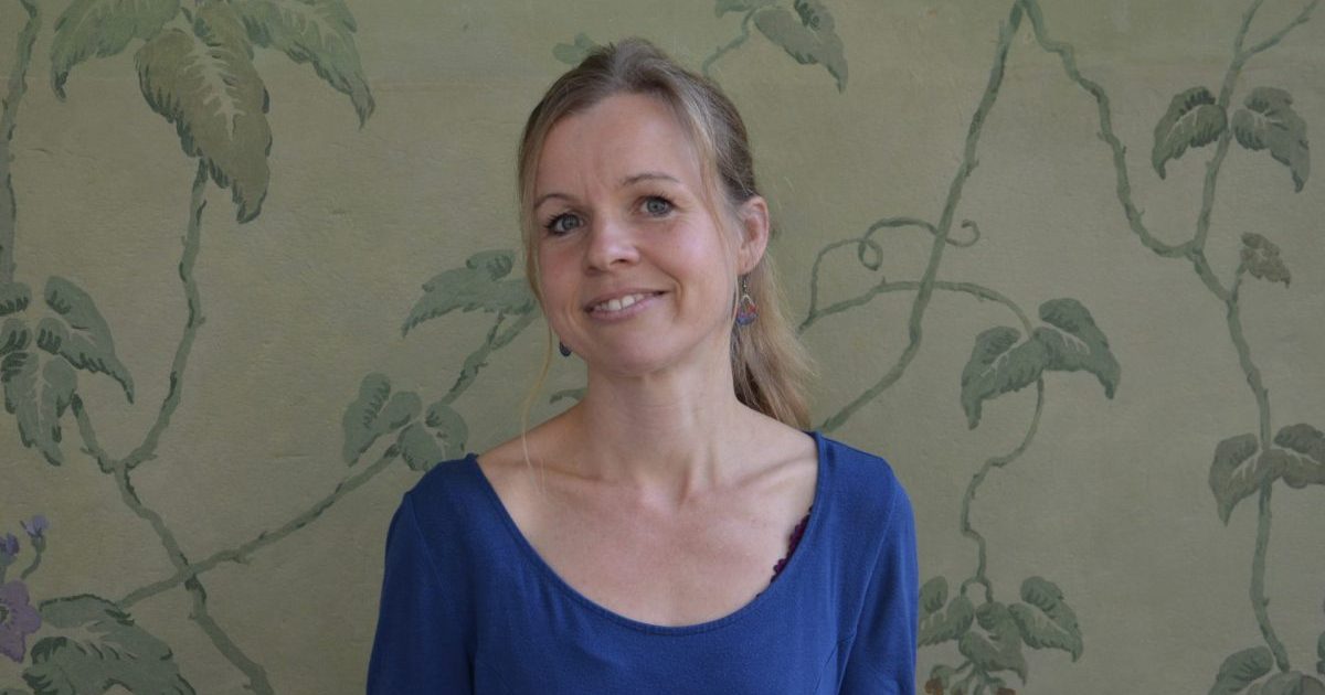 Eine blonde Frau mit zurückgebundenen Haar und blauem Shirt steht vor einer grünlichen Mustertapete.