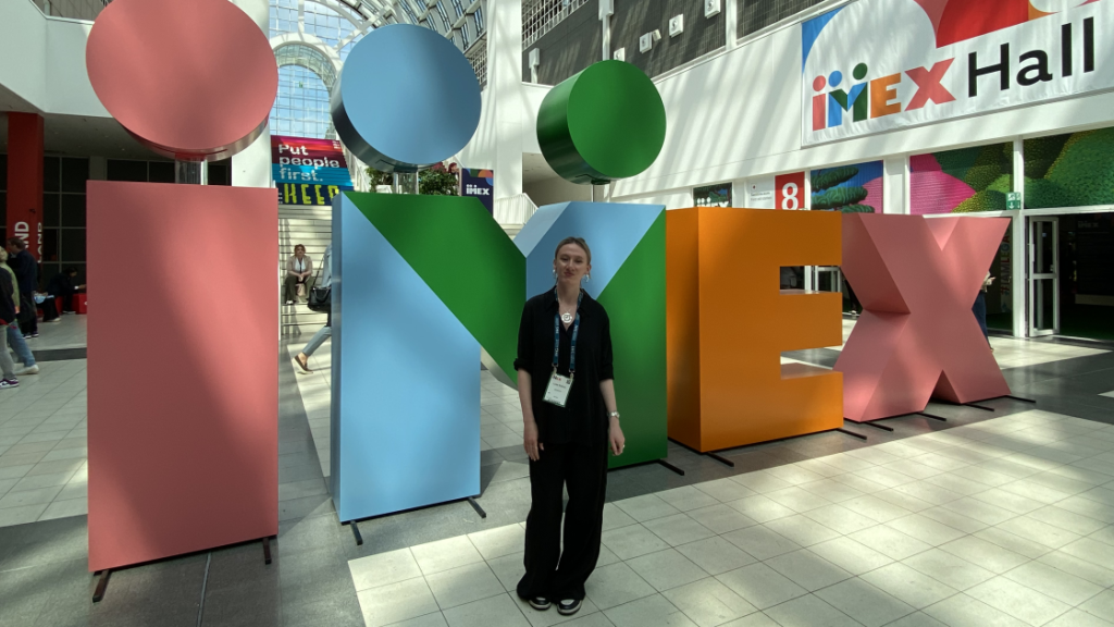 Die JenaKultur-Auszubildende Luise Büttner vor dem IMEX Logo auf der Messe