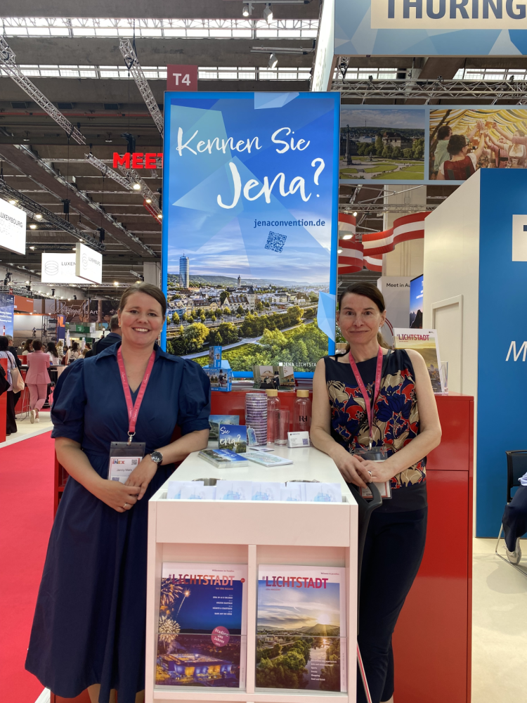 Jenny Marx und Barbara Lütz-Kranich stehen neben dem Stand des Jena Convention Bureaus auf der Messe IMEX.