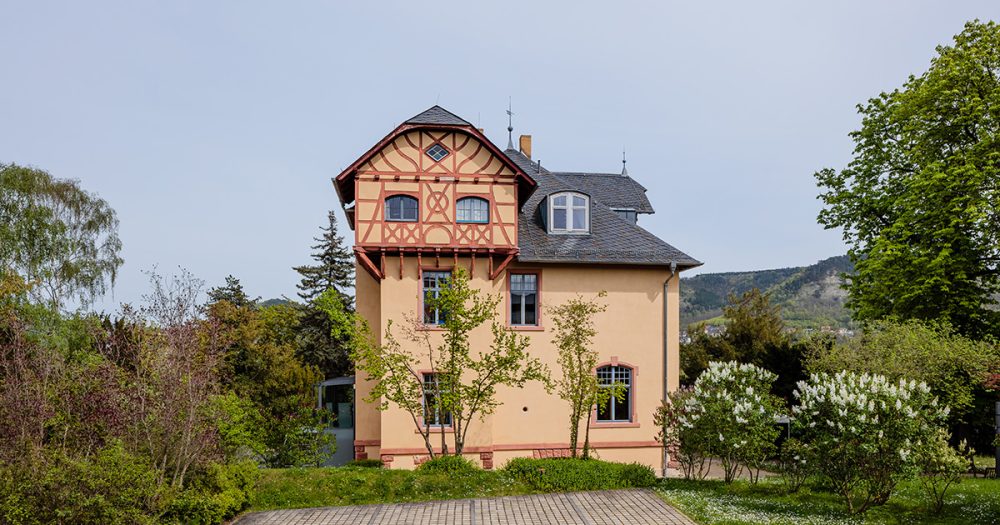 Eine herrschaftliche Villa mit Fachwerk an einem abgesetzten Treppenhaus in einen grünen Garten eingefasst.