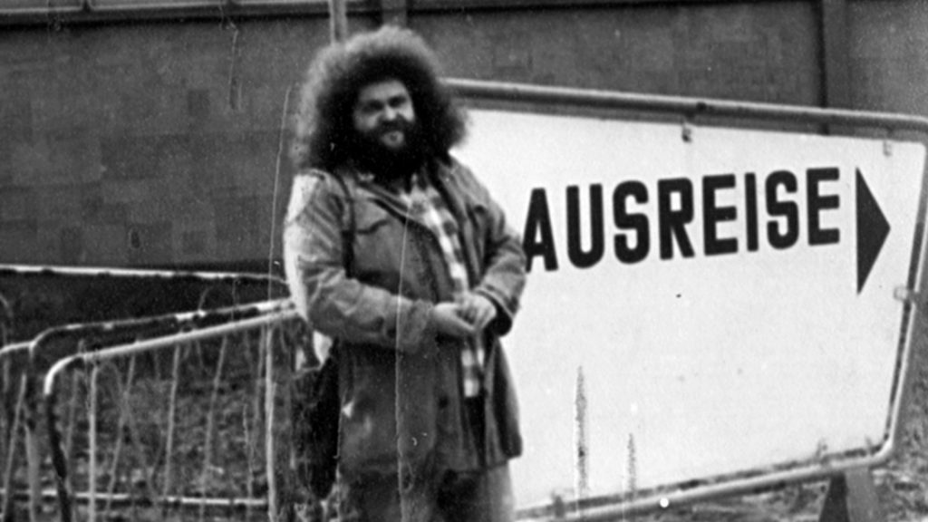 Schwarz-weiß-Fotografie aus DDR-Zeiten. Ein Mann steht mit einem Koffer stolz vor einem Schild mit "AUSREISE" und Pfeil