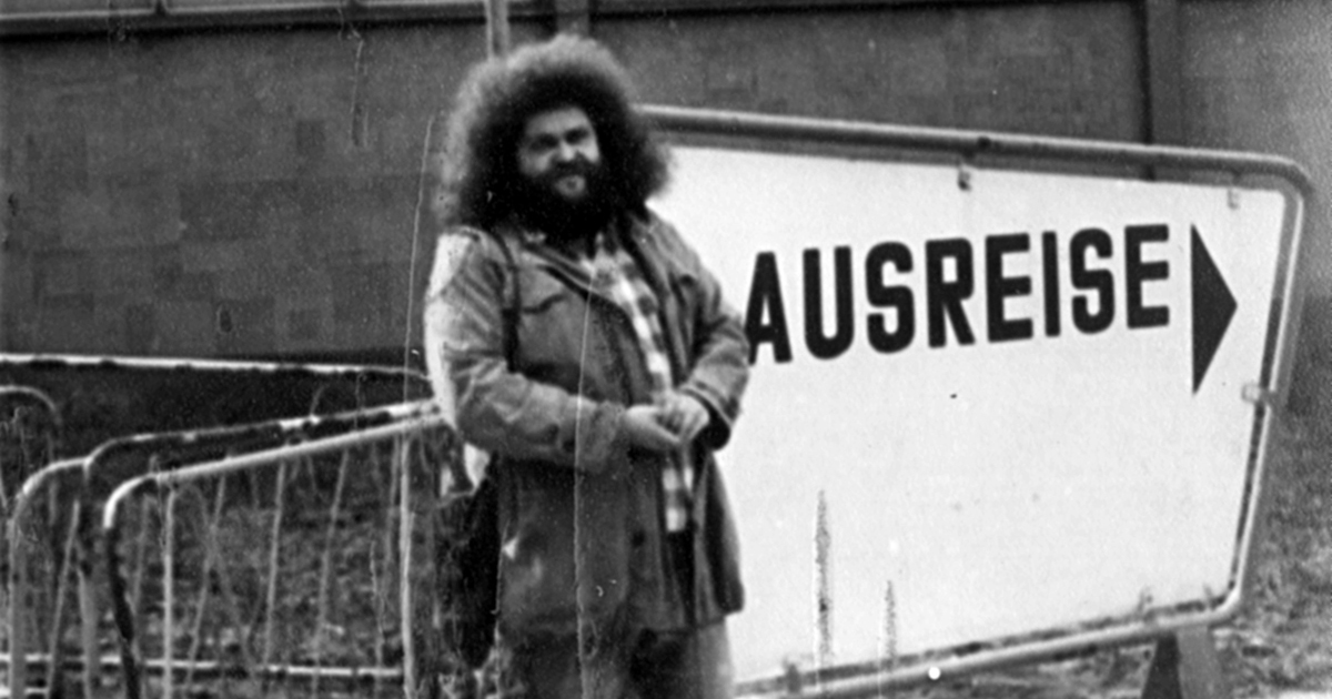 Schwarz-weiß-Fotografie aus DDR-Zeiten. Ein Mann steht mit einem Koffer stolz vor einem Schild mit "AUSREISE" und Pfeil