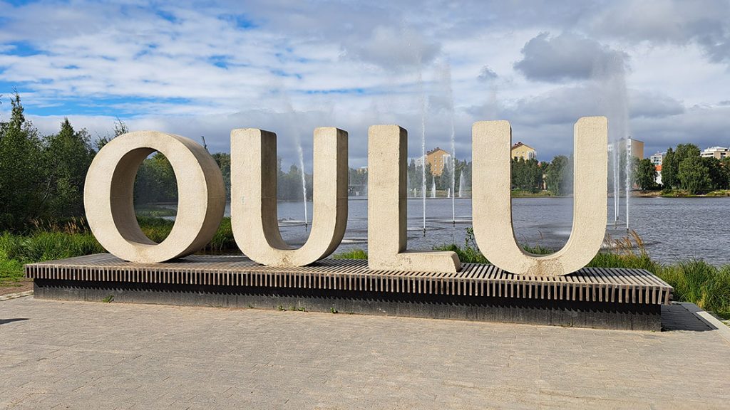 Der Schriftzug "Oulu" in großen Betonbuchstaben vor einem See mit Fontainen