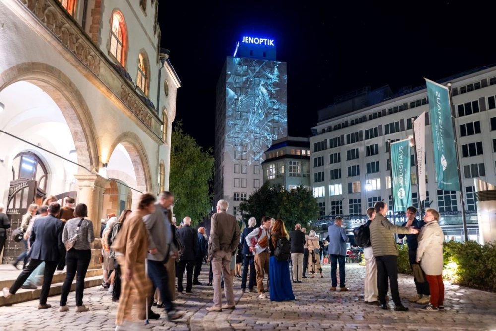 Menschen vor dem Volkshaus Jena, im Hintergrund zu sehen ist das Jenoptik-Gebäude mit Lichtkunst an der Fassade
