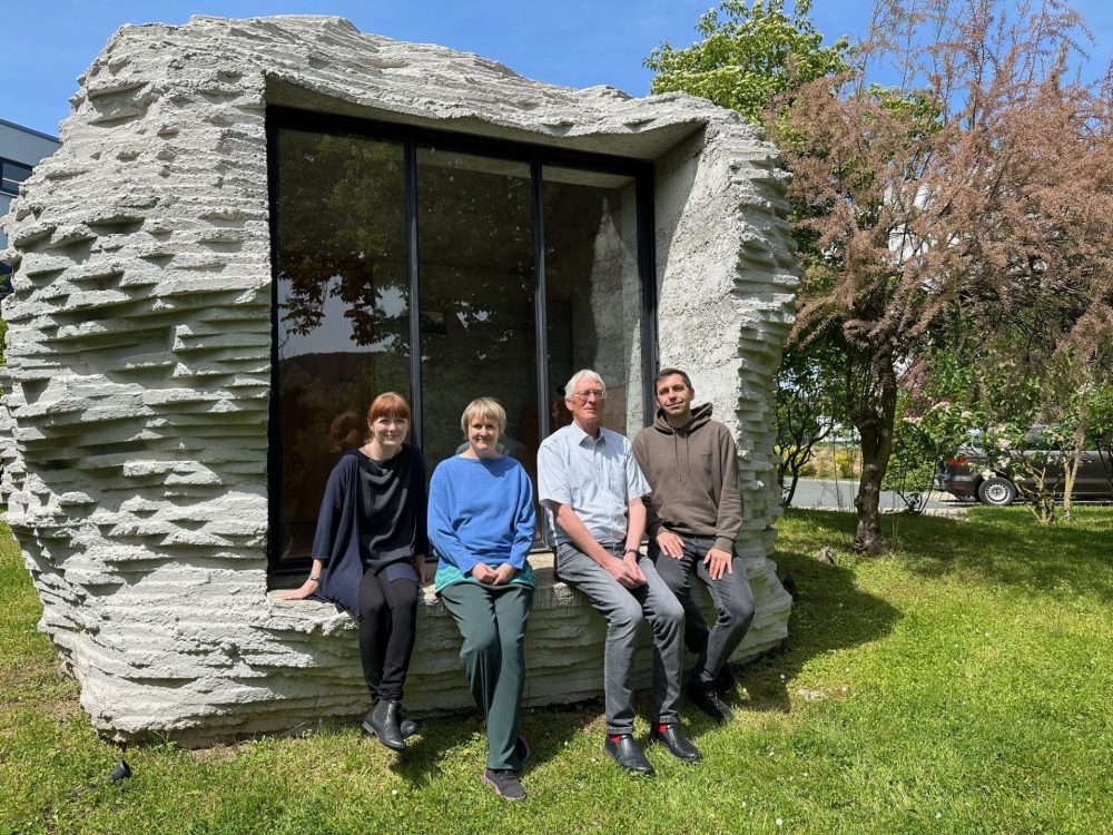 Zwei Frauen und zwei Männer sitzen auf einer steinernen Skulptur mit Fenster in einem Garten und lächeln in die Kamera.