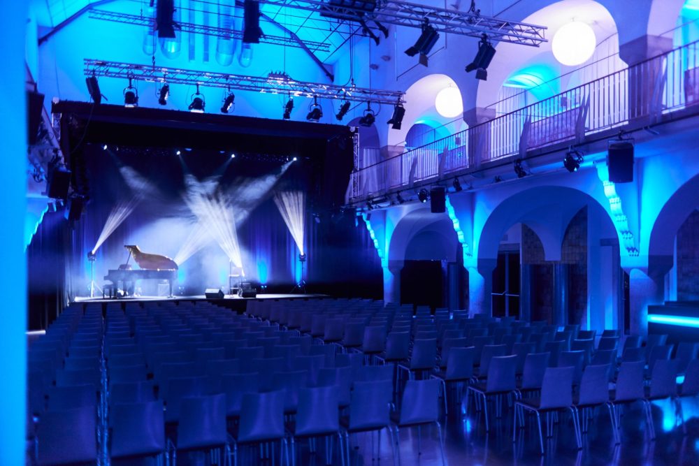 Blau beleuchteter Saal des Volksbad Jena mit Stuhlreihen ohne Gäste, Scheinwerfer strahlen auf die Bühne.