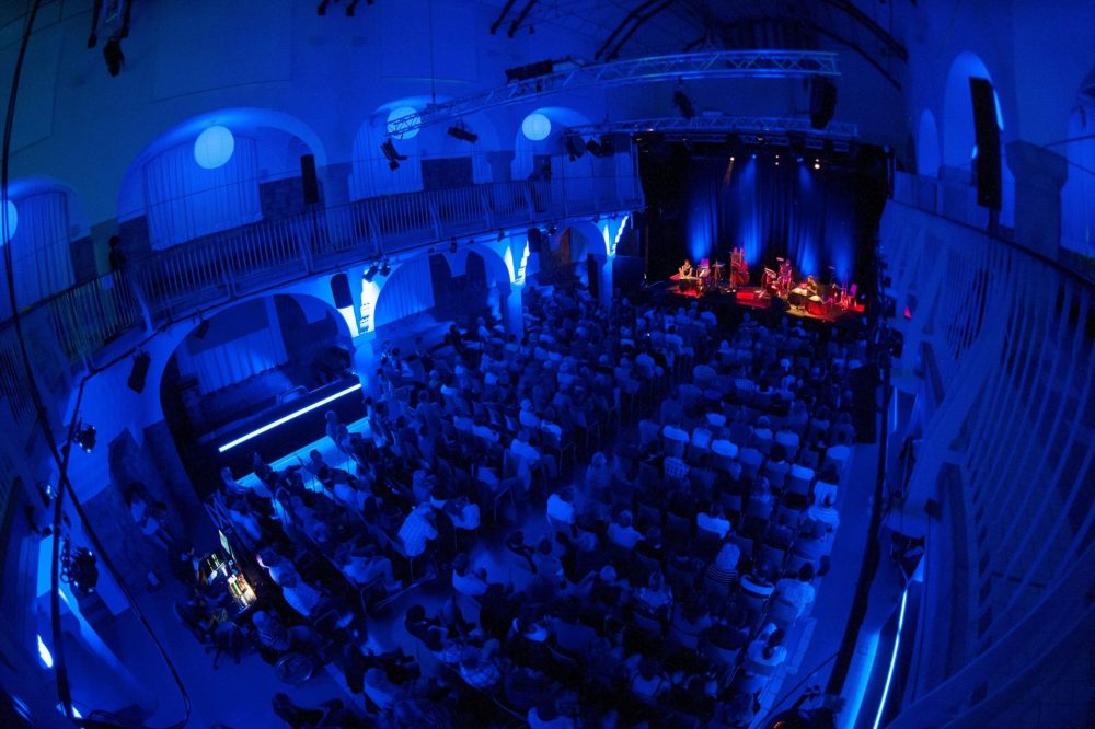 Von Lichtern blau beleuchteter Saal des Volksbad Jena während eines Konzertes, auf den Stühlen sitzen Gäste.