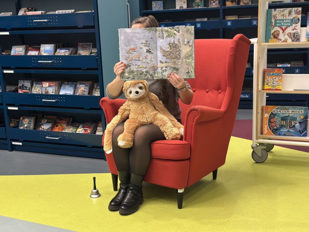 Vorlesen zum Bundesweiten Vorlesetag in der EAB Jena, das Faultier (Maskottchen) der EAB Jena hört ebenfalls gespannt zu, wie die EAB-Mitarbeiterin auf einem roten Sessel sitzend ein Bilderbuch vorliest