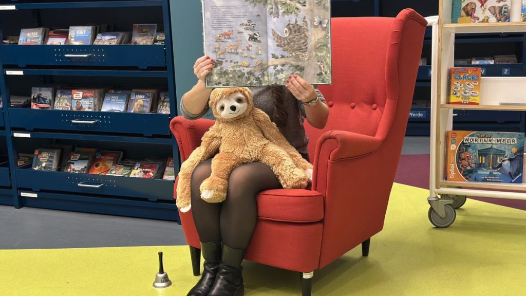 Vorlesen zum Bundesweiten Vorlesetag in der EAB Jena, das Faultier (Maskottchen) der EAB Jena hört ebenfalls gespannt zu, wie die EAB-Mitarbeiterin auf einem roten Sessel sitzend ein Bilderbuch vorliest