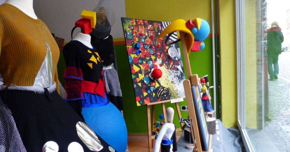 Ein Schaufenster mit Ausstellungsstücken (Kleidung, Gemälde, Holzfiguren etc). und ein kleiner Ausschnitt auf den davor liegenden Gehweg mit Passanten