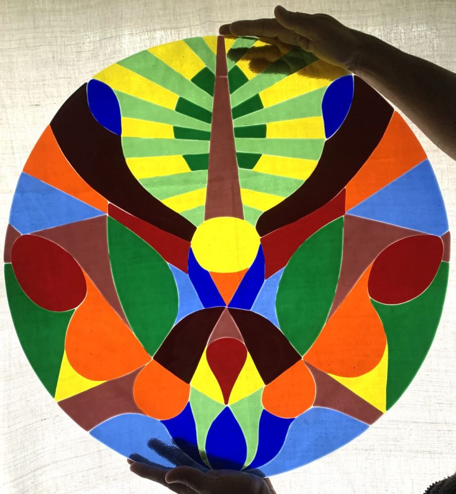 Das fertige Fesnterbild auf Glas. Es ist rund und besteht aus bunten Farben. Es wird oben und unten von einer Hand gehalten.