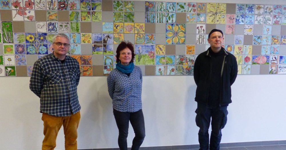 Zu sehen sind die Künstler:innen F. Steenbeck, P. Lieberenz und E. Engelmann. Sie stehen vor dem einer buntem Fliesenprojekt an der Wand im Justizzentrum
