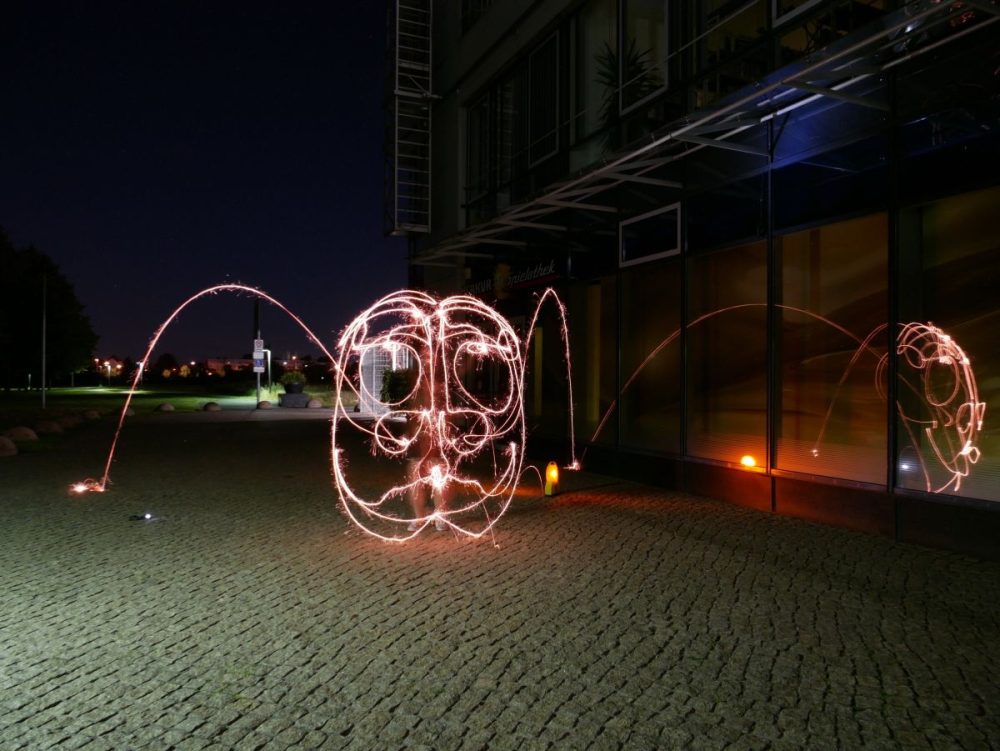 Zu sehen ist ein Bild aus Licht, das durch eine Langzeitbelichtung entstanden ist. Das abstrakte Gesicht ist mit rötlichen Lichtlinien entstanden.