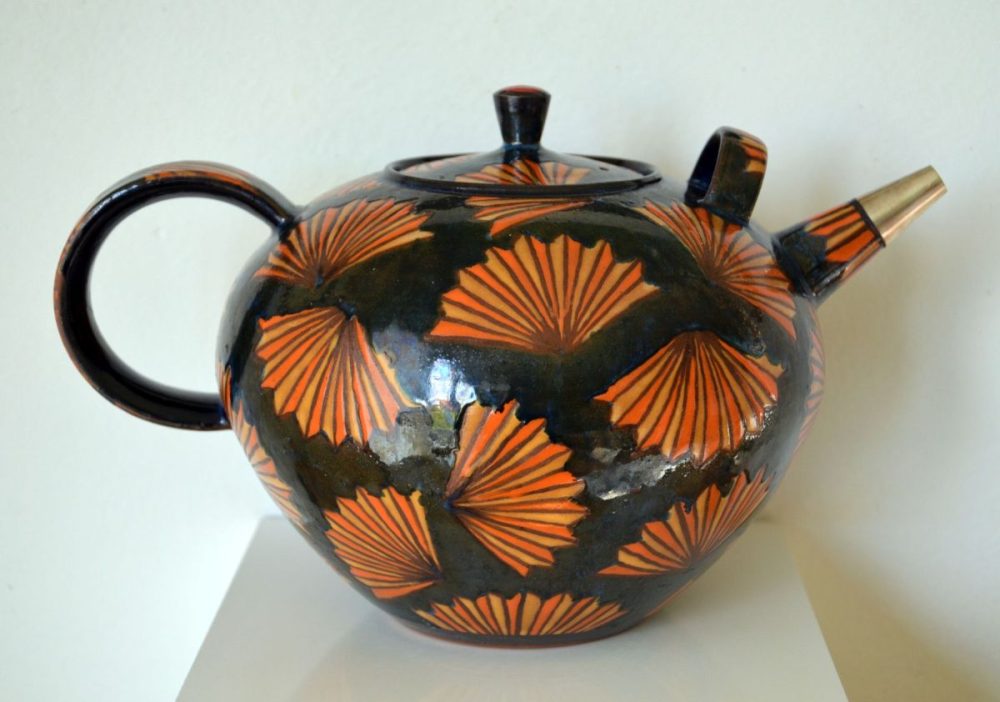 Auf der schwarzen Teekanne sind viele orangfarbene fächer-ähnliche Motive. Die Teekanne hat einen runden Griif und eine goldene Spitze.