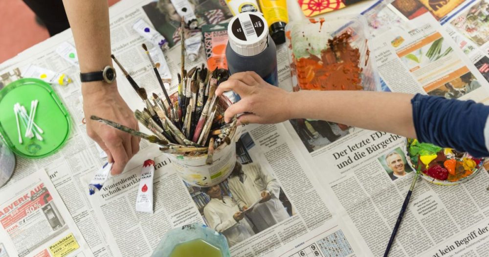 Zu sehen ist ein Tisch, auf dem Zeitungen als Fabrschutz ausgelegt sind. Außerdem ein Becher mit vielen Pinseln und eine Kinderhand, die danach greift.