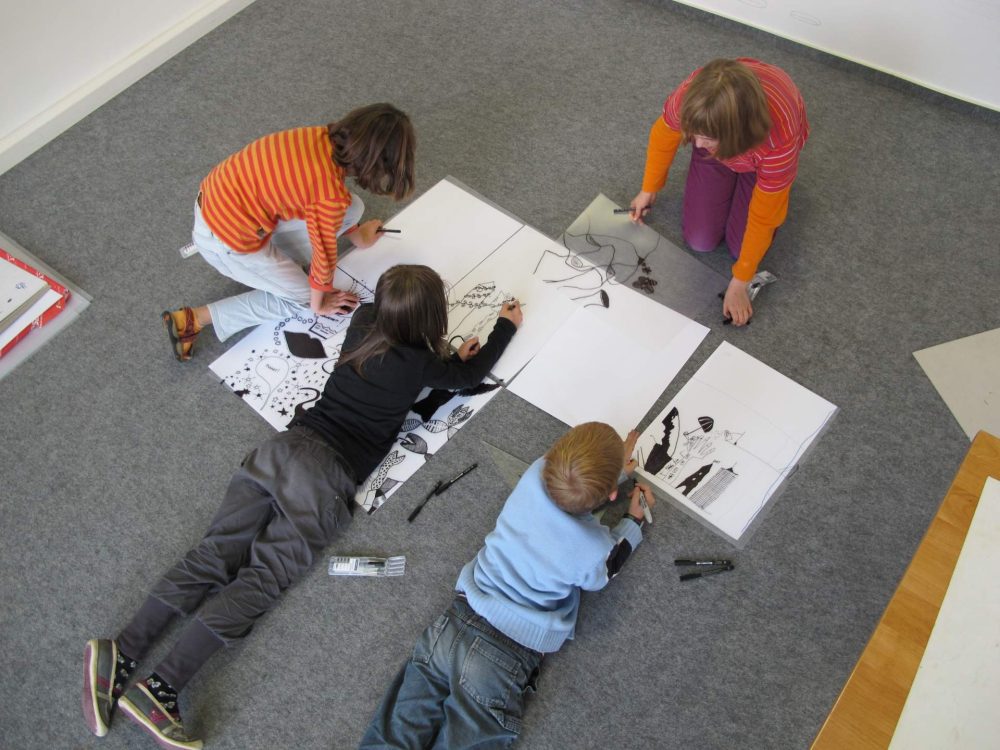 Zu sehen sind vier Schüler:innen, die auf dem Boden Zeichnungen in Schwarz-Weiß auf mehreren A3-Blättern anfertigen.
