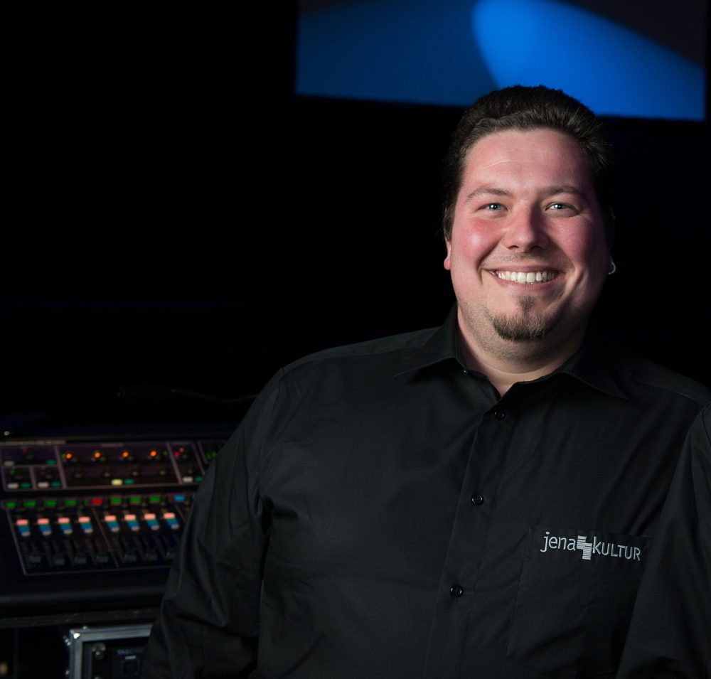 Martin Hädrich in seinen Anfangsjahren bei JenaKultur, er steht vor einem Mischpult und grinst in die Kamera, auf seinem Shirt steht "JenaKultur"