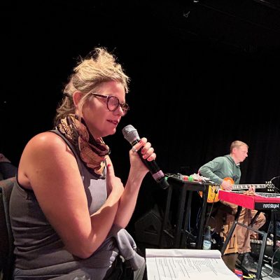 Blick auf die Probebühne im Theaterhaus Jena zur Aufführung des Inklusionsprojektes, im Vodergrund eine Sprecherin, im Hintergrund spiuelt ein Mann Gitarre