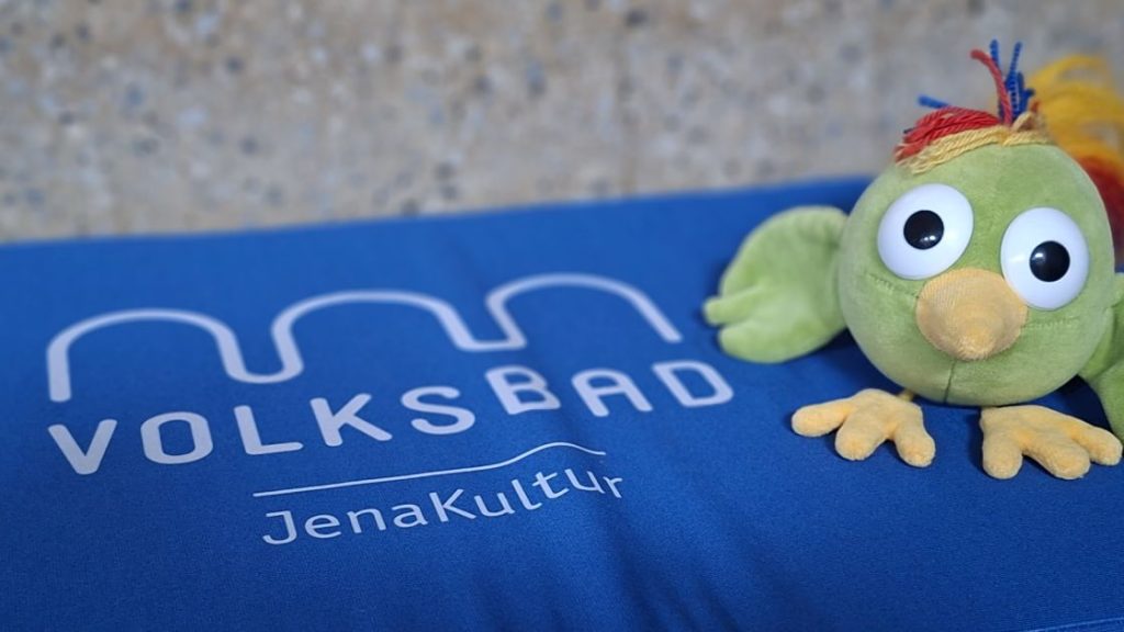 Kuscheltiervogel Adam auf einem blauen Kissen, dass die Ausschrift "Volksbad JenaKultur" hat