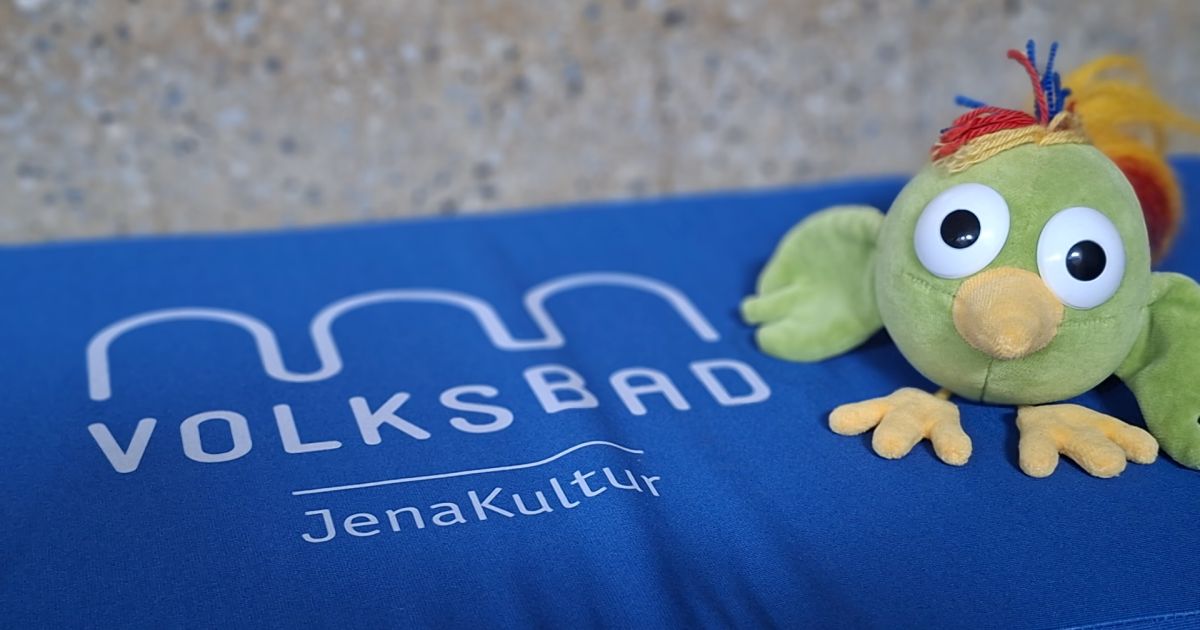 Kuscheltiervogel Adam auf einem blauen Kissen, dass die Ausschrift "Volksbad JenaKultur" hat