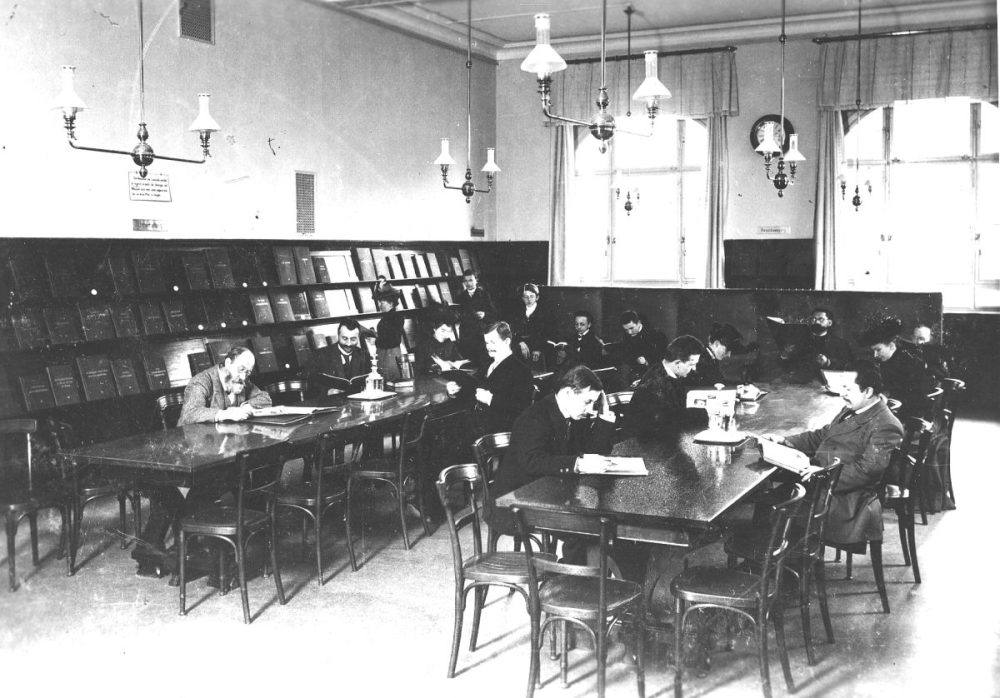 Ein schwarz-weiß-Foto. Zu sehen ist eine große Halle mit holzvertäfelten Wände und schmalen Kronleuchtern. Männer sitzen an Tischen und lesen.