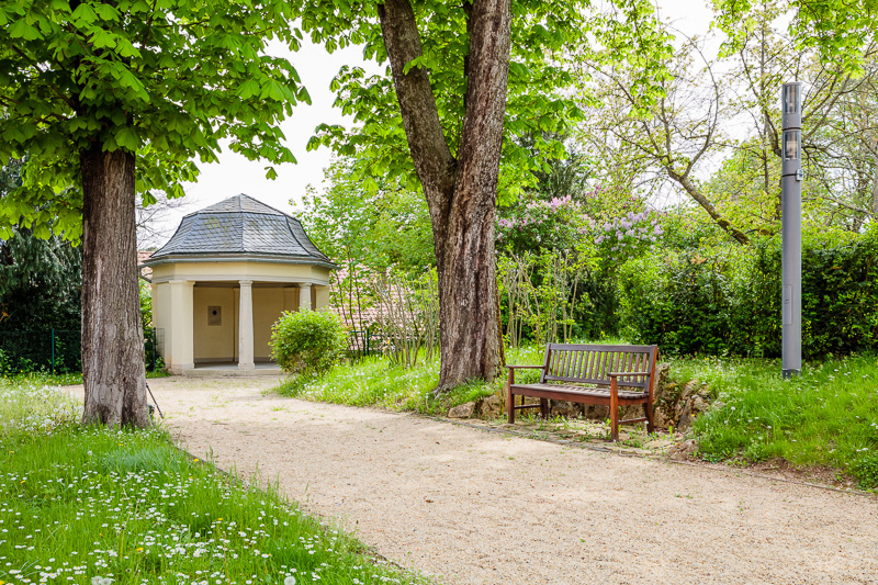 Ein Steinpavillon in einem Garten. Ein Weg führt zum Pavillion, am Wegrand steht eine Bank
