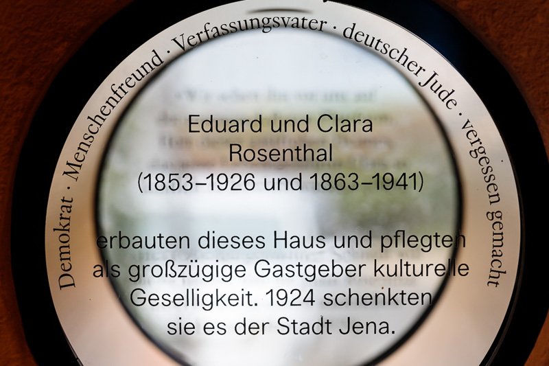 Eine runde milchglasige Scheibe in einer Hauswand. Auf ihr stehen Name und Lebensdaten von Eduard und Clara Rosenthal