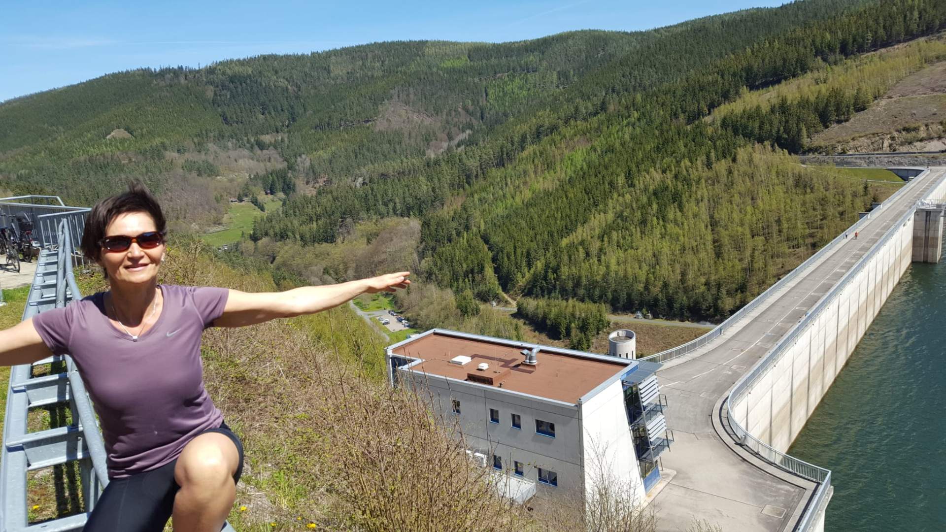 Bild einer Talsperre im Grünen. Links im Bild ist eine Frau in der hocke und breitet die Arme aus. Sie trägt eine Sonnenbrille.
