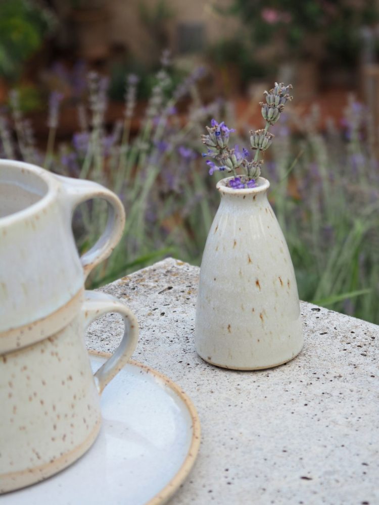 Kleine cremefarbene Keramikvase mit Lavendelzweigen neben Tasse und Teller im Garten.
