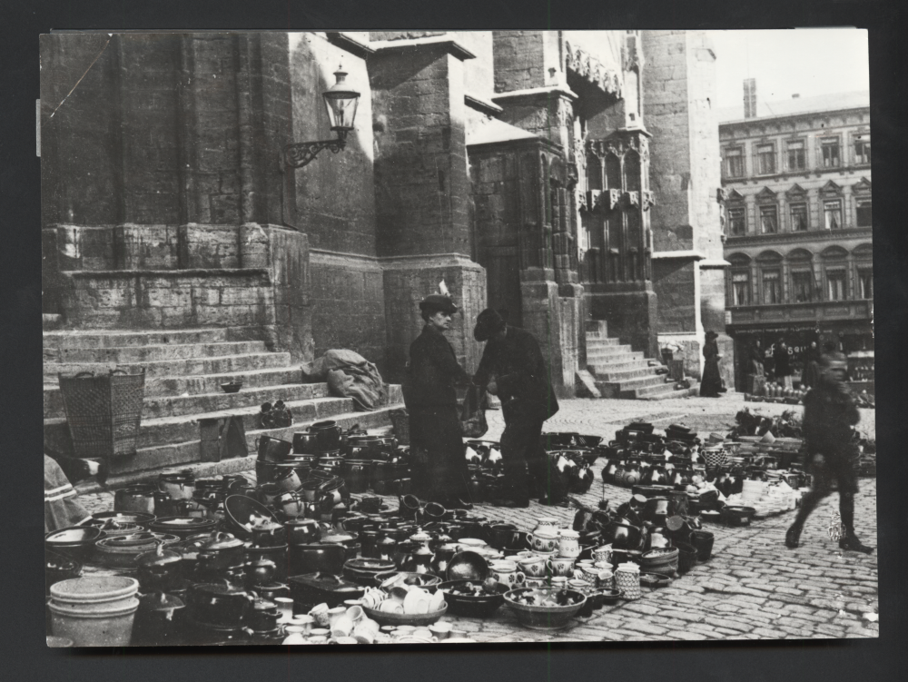 Zwei Männer stehen zwischen vielen Töpfen und Krügen auf einem Markt vor einer Kirche, im Hintergrund Häuserfassaden.