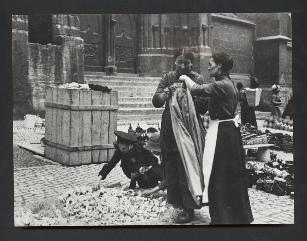 Zwei Frauen und ein Kind stöbern an einem Straßenmarkt vor einer Kirche zwischen Keramik und Haushaltswaren.