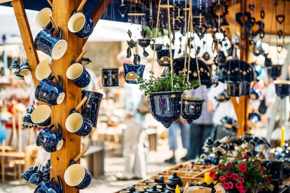 Marktstand mit dunkelblauen Keramiktassen und Blumentöpfen, die an Holzbalken hängen, umgeben von Besuchern.