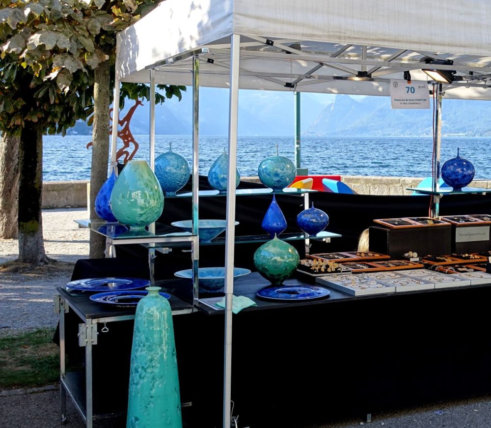Marktstand mit blauen Keramikvasen und Schalen am Seeufer, im Hintergrund Wasser und Berge.