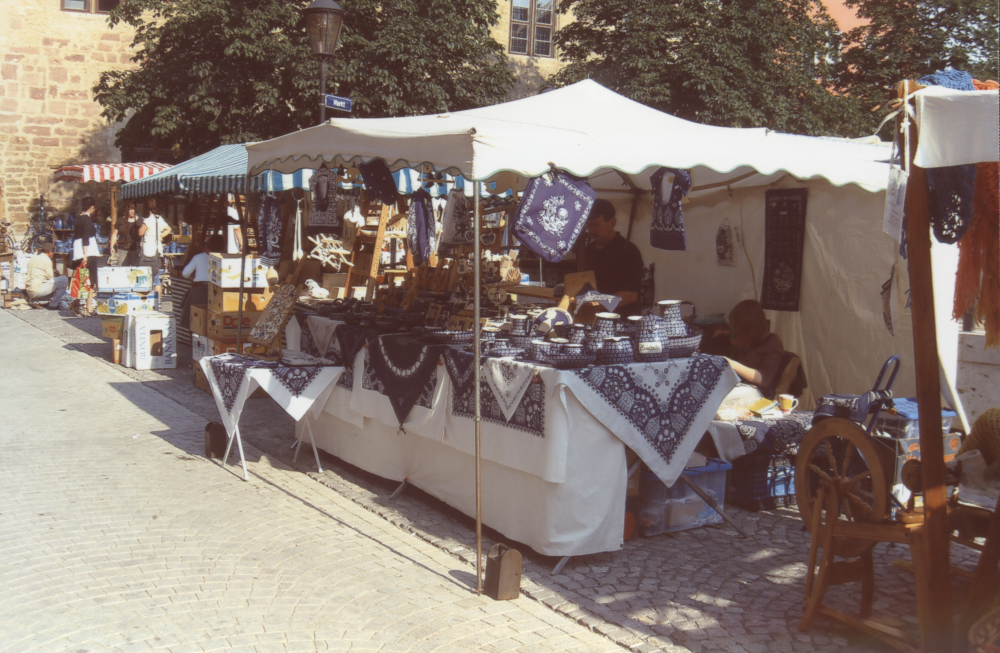 Marktstand mit blau-weißer Keramik und Textilien, Verkäufer bedient Kunden, weitere Stände im Hintergrund.