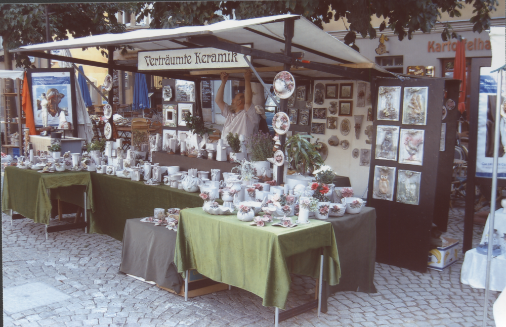 Marktstand „Verträumte Keramik“ mit vielen bemalten Keramiken auf grünen Tüchern, Händler richtet das Dach.