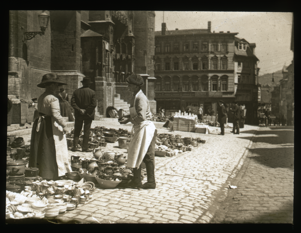 Straßenmarkt mit Keramikgeschirr neben einer Kirche, mehrere Personen in historischer Kleidung auf dem Kopfsteinpflaster.