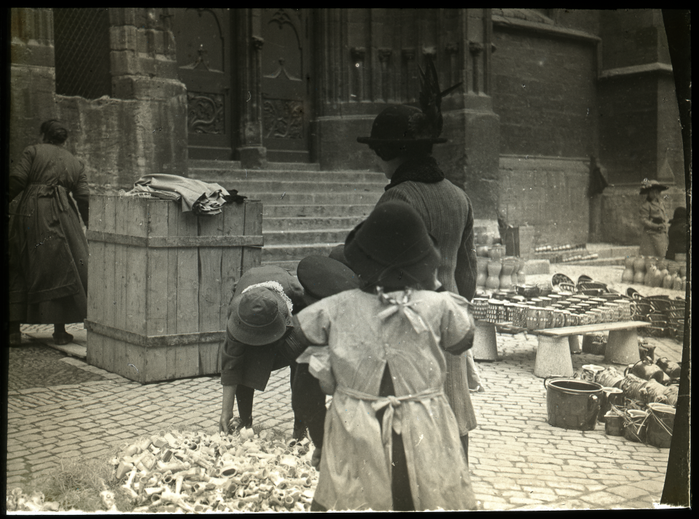 Eine Frau mit zwei Kindern betrachtet Waren auf einem Straßenmarkt vor einer Kirche, Keramikgefäße liegen am Boden.