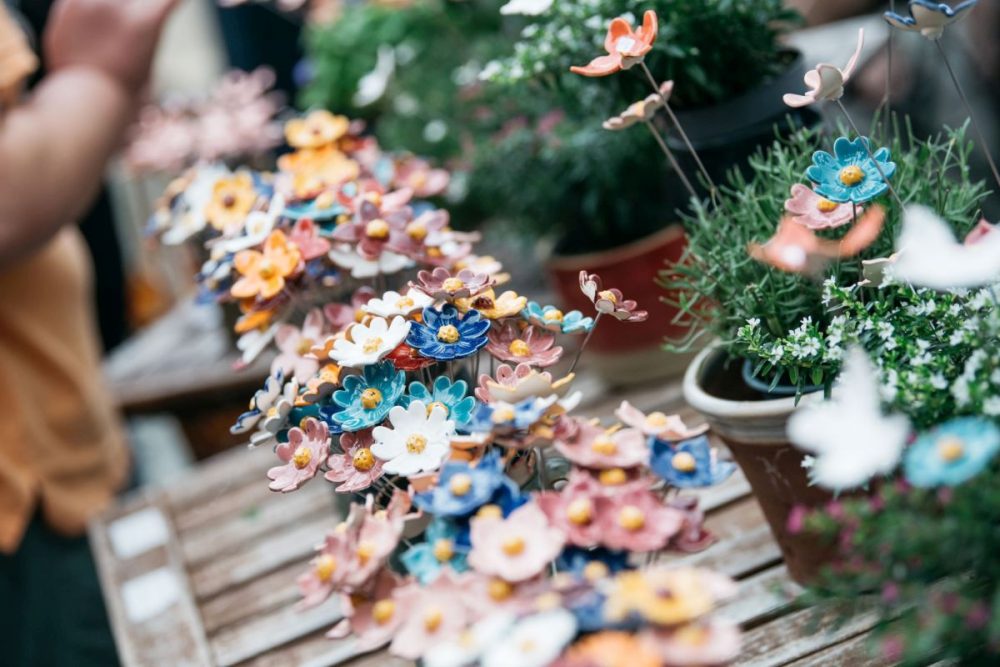 Bunte Keramikblumen in verschiedenen Farben zwischen grünen Topfpflanzen auf einem Holztisch.