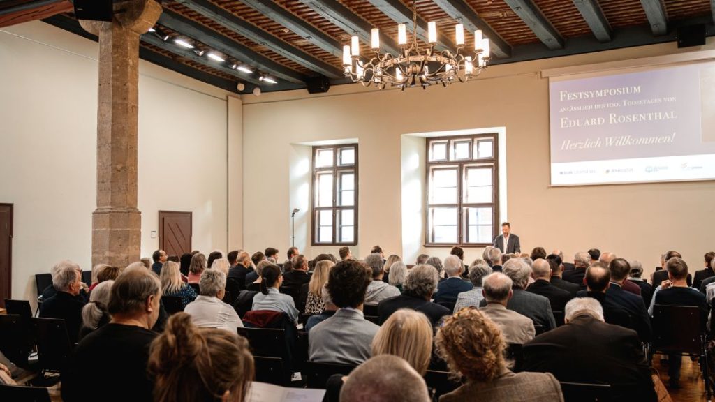 Mann im Anzug spricht am Rednerpult, vor ihm sitzendes Publikum im Saal.