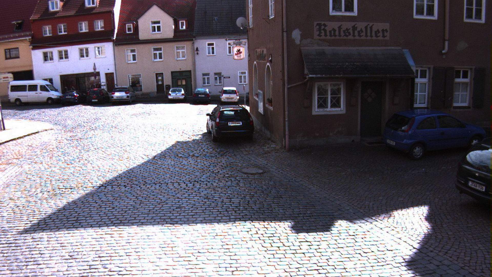 Der Stadthof in Jena Lobeda-Altstadt vor der Baumaßnahme, die Straße ist mit Kopfsteinpflaster ausgelegt. Im Vordergrund ein parkendes Auto.